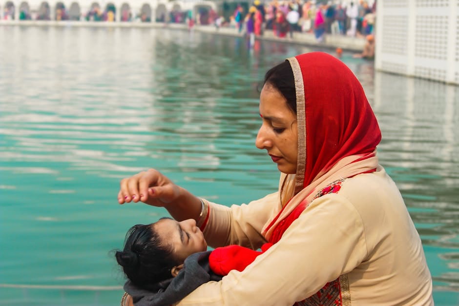 A mother comforts her child by the serene waters, wearing a red headscarf.