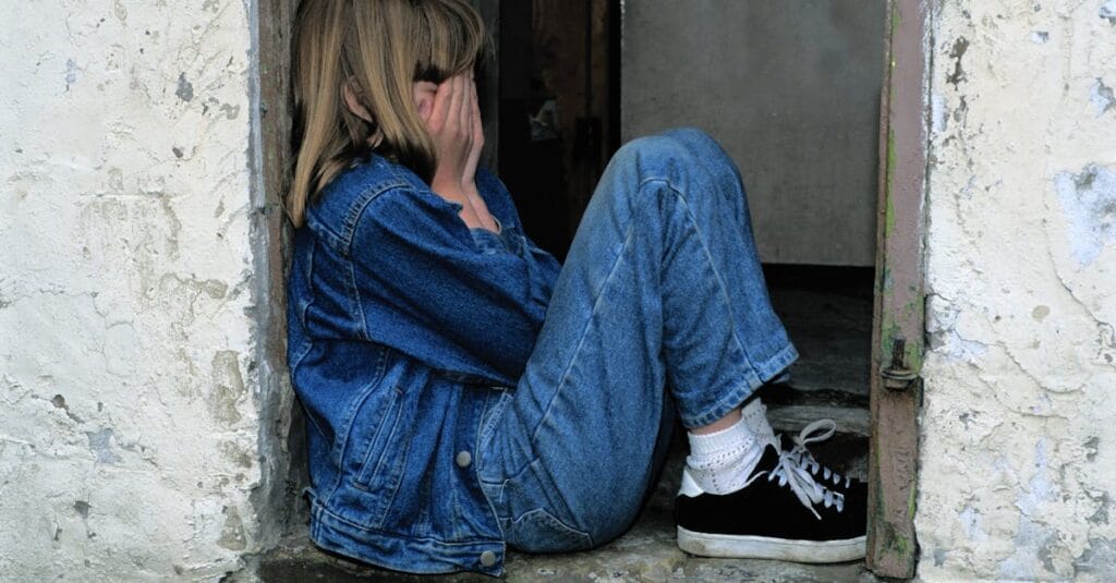 A young girl in denim sits alone in a doorway looking distressed, conveying loneliness.