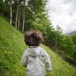 A young boy wearing a hoodie runs through a lush, mountainous landscape filled with trees and wildflowers.