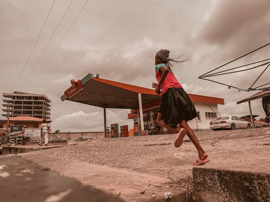 A young girl runs past a gasoline station under a cloudy sky in an urban environment.