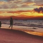Silhouettes of a child and adult running along a beach at sunset with a dramatic sky and gentle waves.