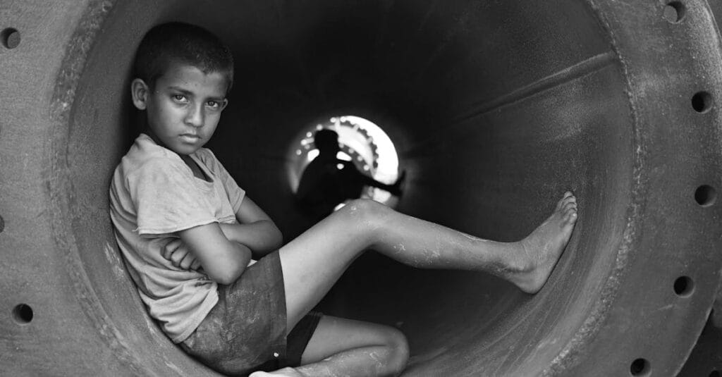 A contemplative young boy sits barefoot in a large industrial pipe in black and white.