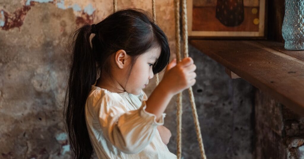 Thoughtful young girl sitting on a swing indoors, in a rustic setting with soft lighting.