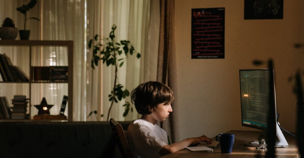 A young boy intensely focused on coding on a computer in a cozy, dimly-lit room.