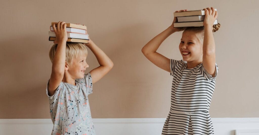 Two smiling children balancing books on their heads indoors, symbolizing fun in learning.