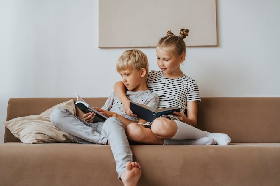 Two siblings sitting on a sofa reading books together indoors.