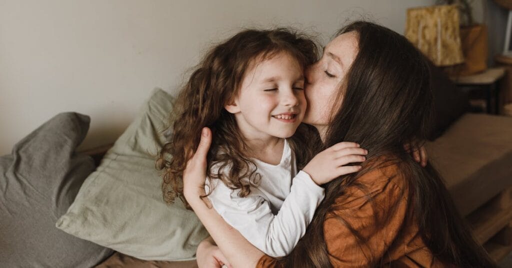 A touching moment of a mother kissing her daughter indoors, radiating love and warmth.
