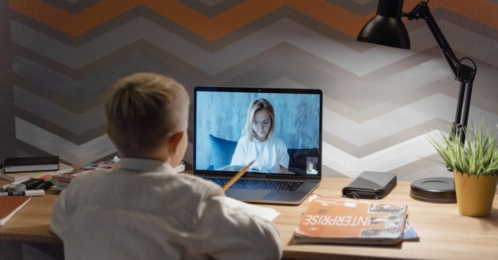 A child participates in an online lesson from home, using a laptop for video conferencing with a teacher.
