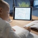 A young boy focuses on math problems on a tablet while studying at a desk indoors.