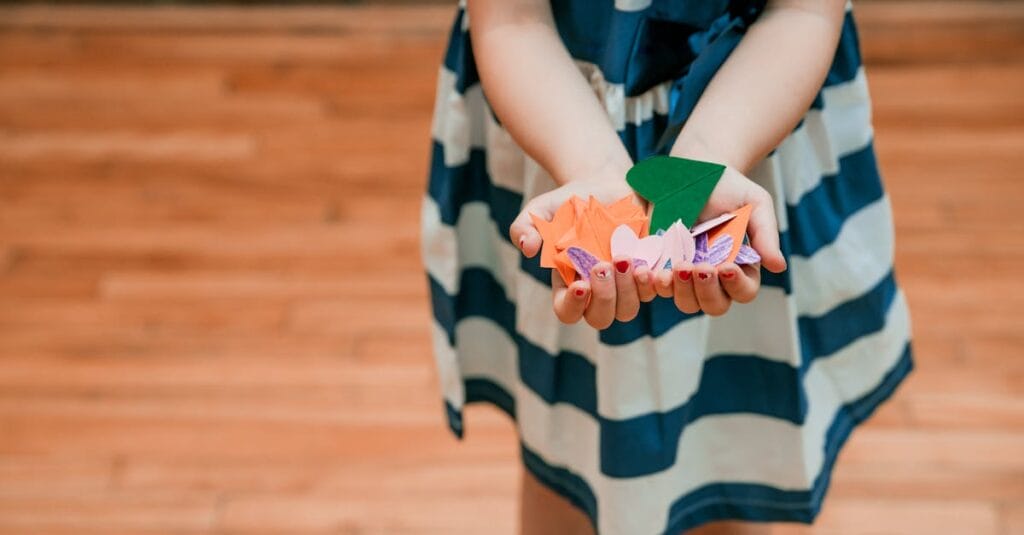 Child displaying colorful paper origami shapes in hands, indoors.