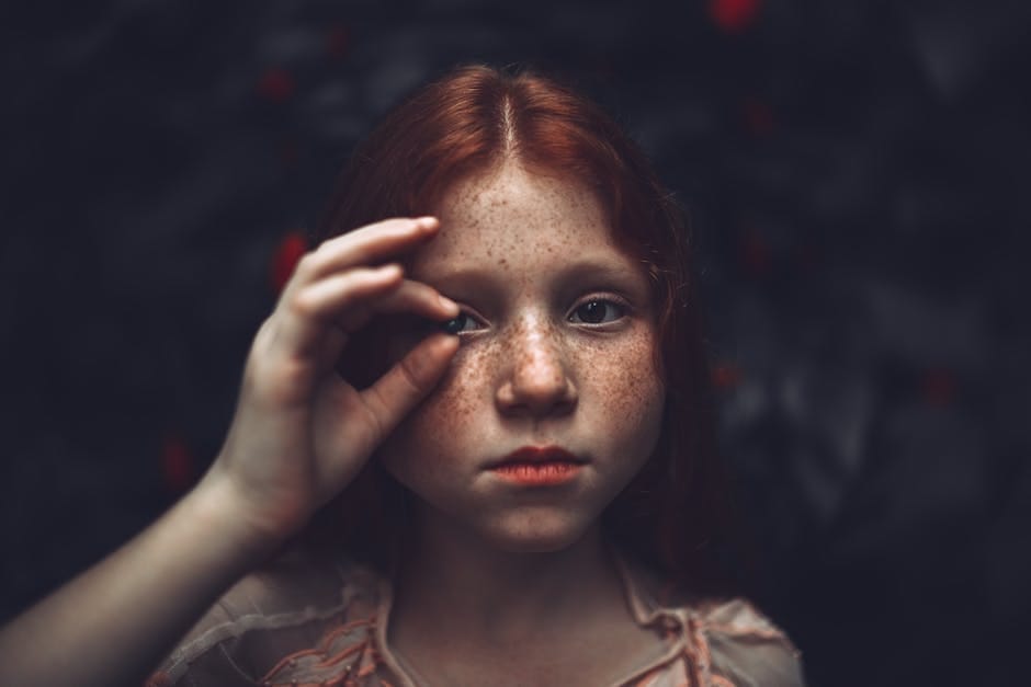 A striking close-up portrait of a young freckled redhead girl holding a pebble to her eye with a mysterious expression.
