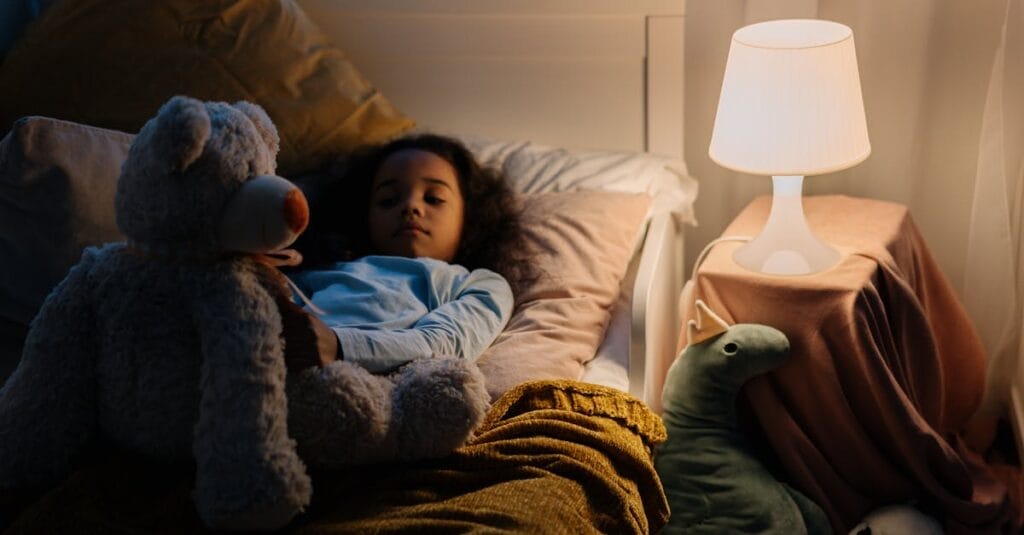 Young girl resting in bed with a teddy bear and toys, illuminated by a bedside lamp.