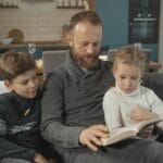 A father reading a book with his two children on a sofa in a warm indoor setting.