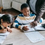 High angle of crop unrecognizable black female teacher explaining task to focused little schoolboy sitting at desk near attentive Asian classmate writing in notebook