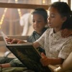 Two siblings enjoying a storybook together, surrounded by cozy lighting indoors.