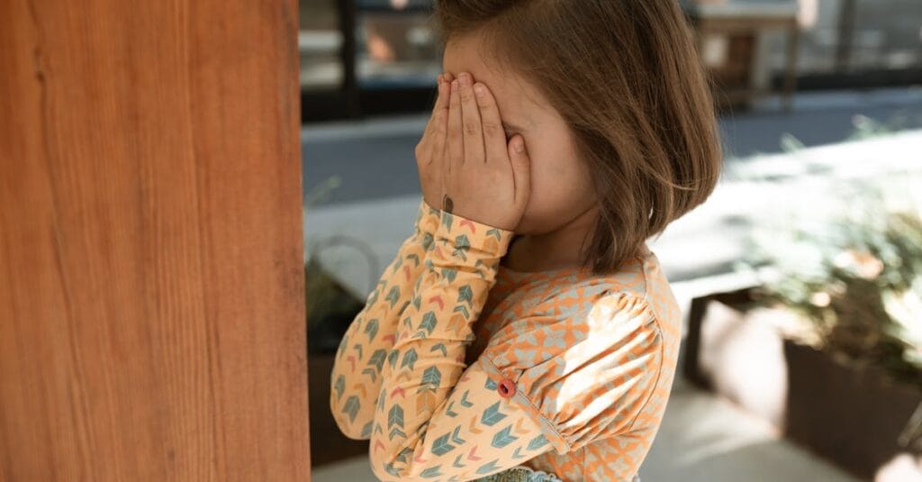 A shy child with brunette hair covers her face while standing outdoors in a playful moment.