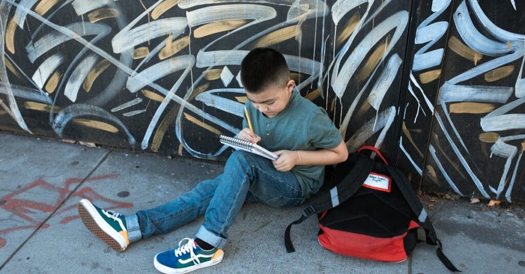 Child sitting on sidewalk writing in a notebook beside graffiti wall with a backpack.