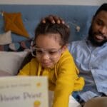 A father and daughter share a joyful moment reading on the bed.
