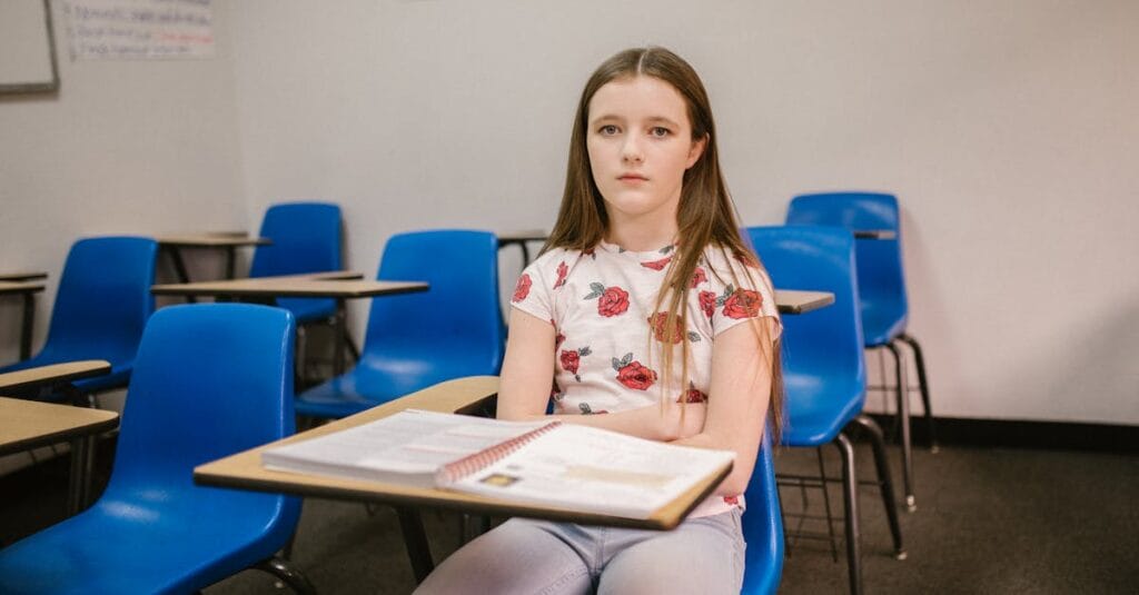 A young girl sits alone in an empty classroom looking pensive and isolated.