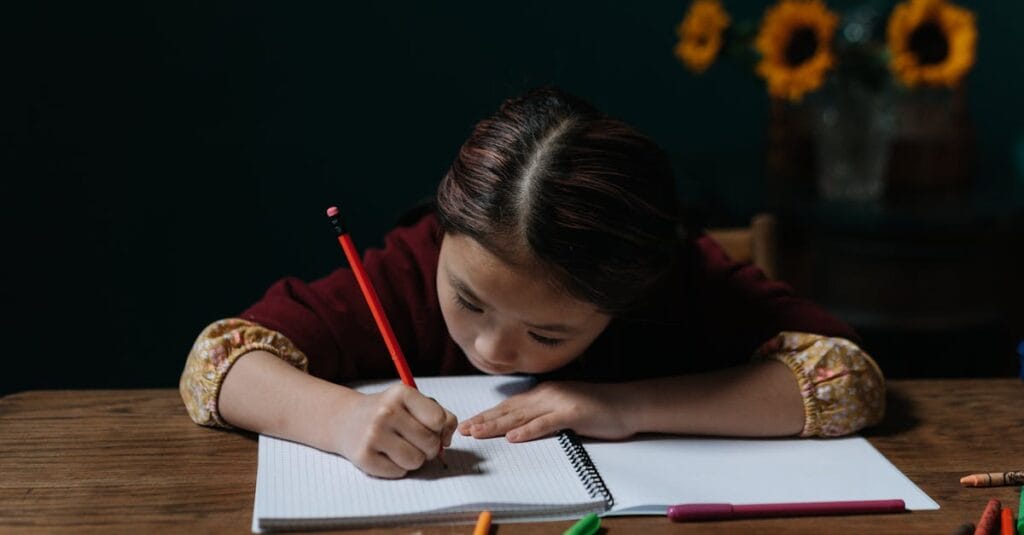 An East Asian child writing in a notebook at a wooden table, focused on homework indoors.