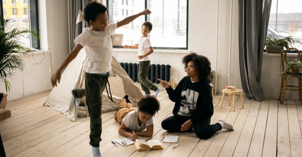 Full body of African American mother sitting on wooden floor near daughter writing in copybook in light room with playing boys