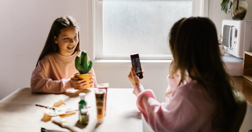 Two girls enjoy arts and crafts indoors, with one capturing the moment with a smartphone.