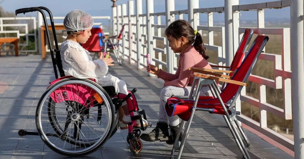 Two young girls enjoying a sunny day on a terrace, one in a wheelchair, sharing a sweet moment.