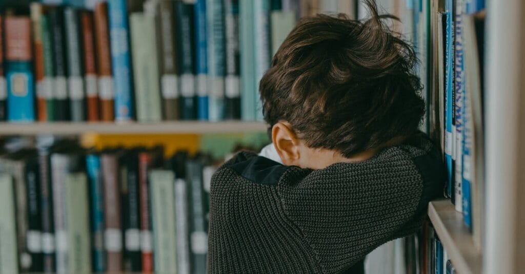 A young boy looking down, leaning against a bookshelf in a library, evoking a sense of loneliness.