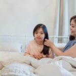 A mother lovingly fixes her daughter's hair on a cozy bed, capturing a moment of bonding and care.