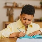 A young boy with curly hair focused on writing in a notebook at home.
