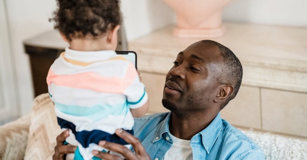 A father holding his young child indoors, sharing a joyful moment at home.