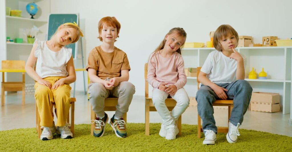 Group of children sitting on chairs, smiling inside a modern classroom setting.