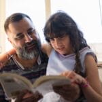 A loving father reads with his daughter by a sunlit window, fostering family bonding.
