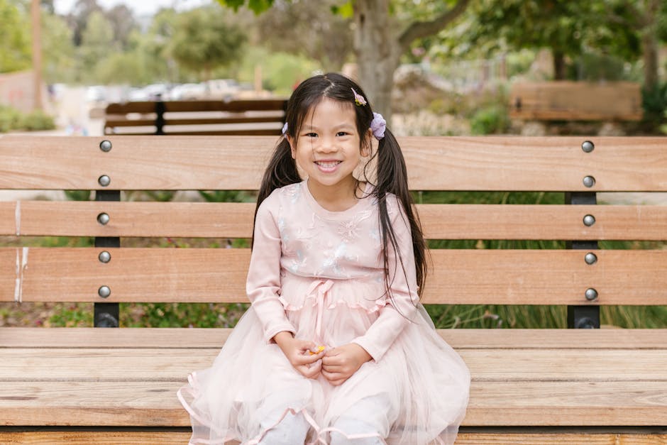 Cheerful young girl in a pink dress smiling while sitting on a park bench outdoors.