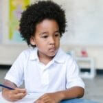 A focused young boy with afro hair writing in a classroom setting, showcasing education and learning.
