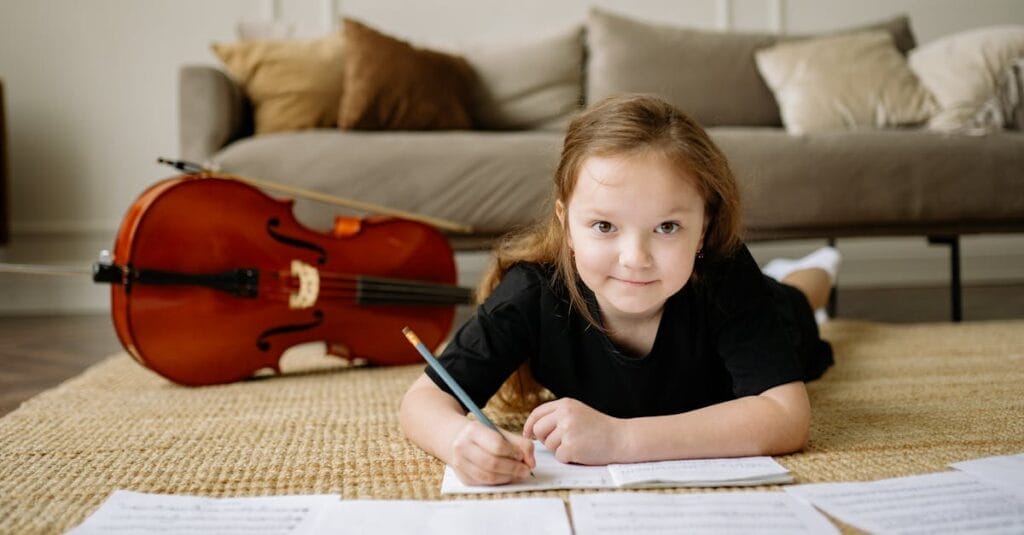 A young girl writing musical notes on the floor next to her cello, creating a cozy home music practice scene.