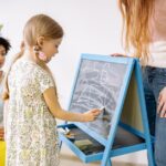 A young girl writes numbers on a chalkboard in a classroom, supervised by adults.