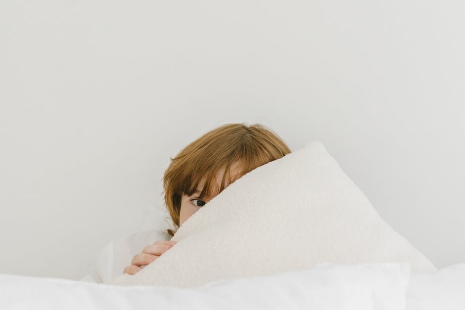 A shy child peeks over a soft white pillow in a cozy indoor setting.