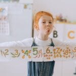 Happy redhead child holding number cards in a classroom, enjoying fun learning.