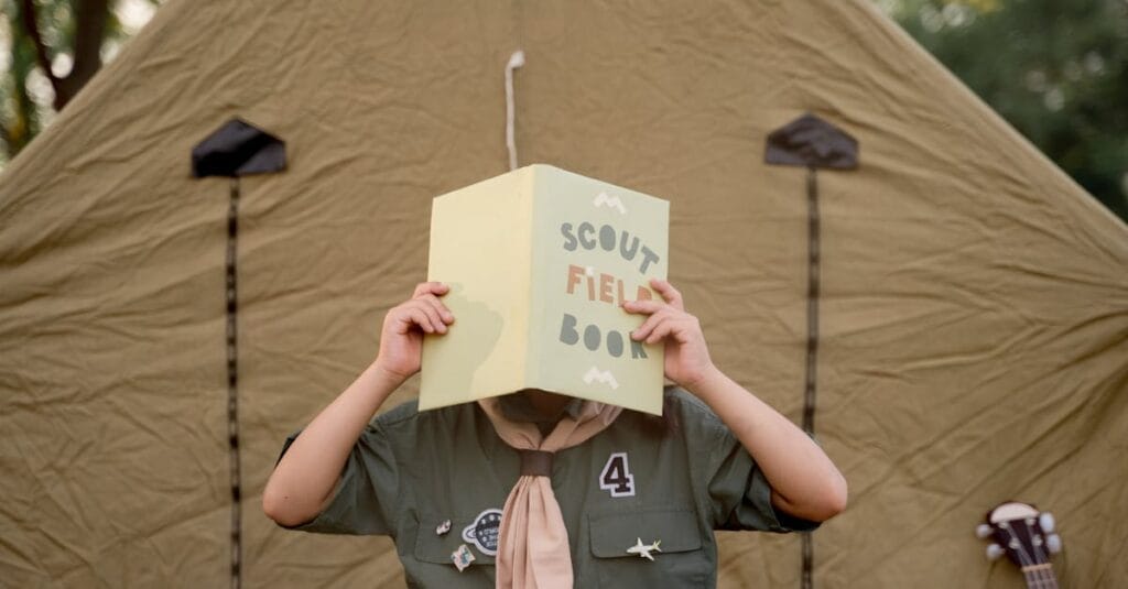 A scout child reads a field book outside a tent, embracing outdoor adventure.