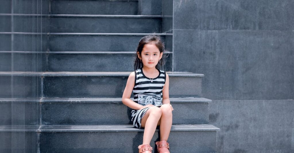 A young girl in a striped dress sits on a sleek grey staircase, looking into the camera.