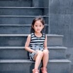 A young girl in a striped dress sits on a sleek grey staircase, looking into the camera.