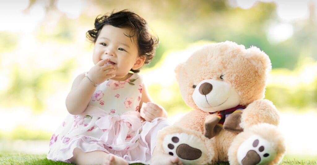 Smiling baby girl sits on grass with a plush teddy bear, enjoying a sunny day.