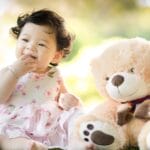 Smiling baby girl sits on grass with a plush teddy bear, enjoying a sunny day.