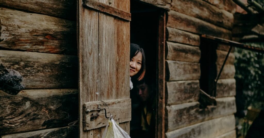 A child peeks playfully from a rustic wooden cabin door on a sunny day.