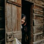 A child peeks playfully from a rustic wooden cabin door on a sunny day.