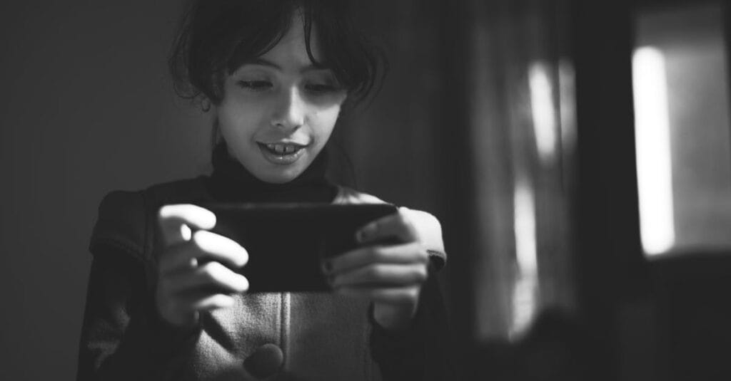 A young girl smiling while using a smartphone in a black and white setting indoors.