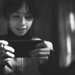 A young girl smiling while using a smartphone in a black and white setting indoors.