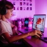 Child focused on laptop wearing headphones at desk with purple lighting. Modern tech scene.