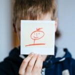A child holding a hand-drawn stick figure portrait indoors.